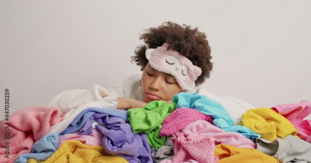 Unhappy tired woman sticks head in pile of multicolored laundry wears sleepmask on forehead overwhelmed by housewotk cleans out wardrobe looks displeased not eager to fold clothes after washing Unhappy tired woman sticks head in pile of multicolored laundry wears sleepmask on forehead overwhelmed by housewotk cleans out wardrobe looks displeased not eager to fold clothes after washing