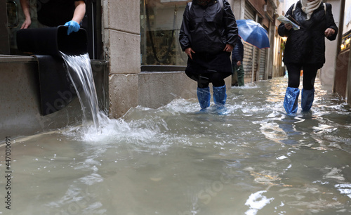 Fototapeta Naklejka Na Ścianę i Meble -  worker empties the water from her shop with a bucket and other people with waterproof gaiters as they walk in the street completely flooded by the high tide in Venice