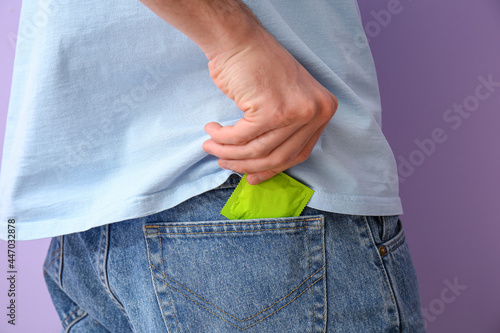 Man putting condom into pocket of jeans on color background, closeup photo