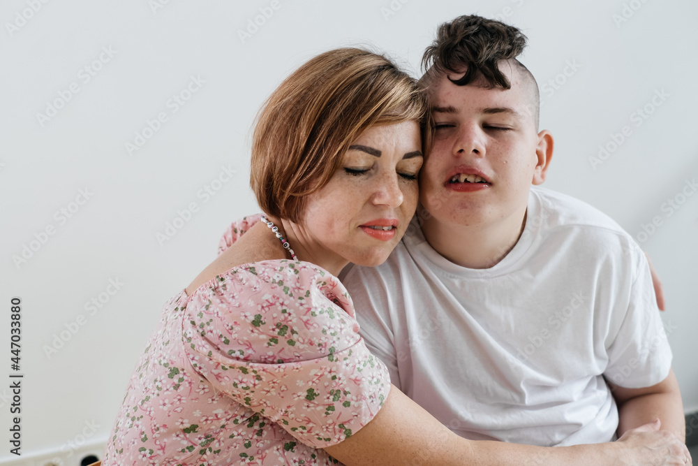 Mother hugging disabled boy 17 years old in wheelchair with cerebral palsy over white background. Woman assists person with Special Needs. Emotional family lives with disability, holding hands Mother hugging disabled boy 17 years old in wheelchair with cerebral palsy over white background. Woman assists person with Special Needs. Emotional family lives with disability, holding hands