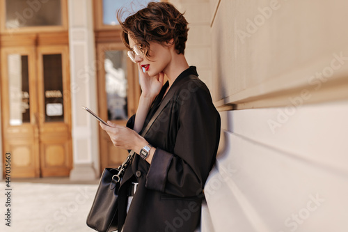 Profile photo of cool girl with short hair in black jacket holds tablet outdoors. Beautifuul woman in eyeglases with handbag posing at street.. photo