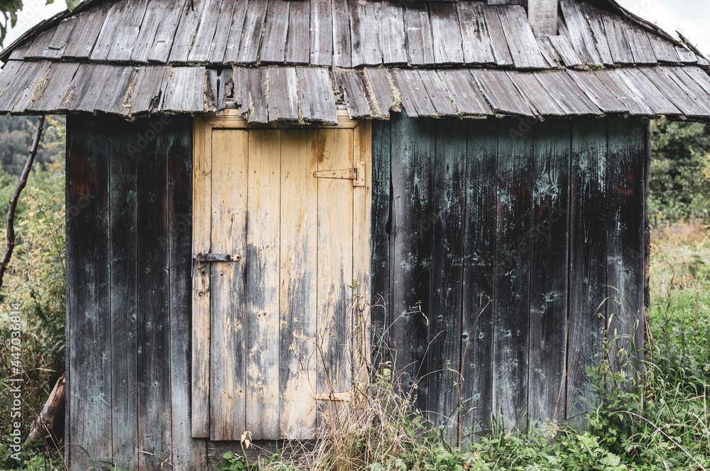 Naklejka premium An old small Carpathian wooden house, blackened with age, with a traditional wooden tile roof. A yellow grunge door with peeling paint is closed with a rusty iron bolt with hinges made by a blacksmith