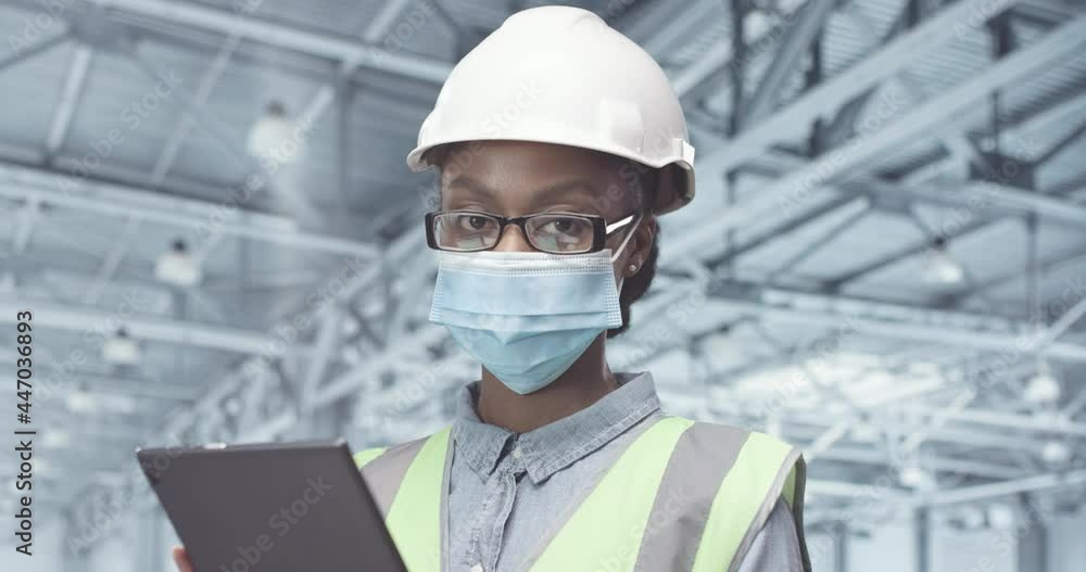 Close up portrait of young african american woman in white helmet and protective medical mask standing at factory with tablet. Close up portrait of young african american woman in white helmet and protective medical mask standing at factory with tablet.