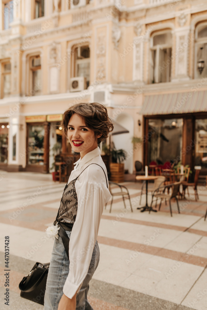 Happy girl in light shirt and jeans looking into camera in city. Modern woman with short hair and bright lips smiling at street..
