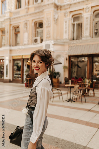 Happy girl in light shirt and jeans looking into camera in city. Modern woman with short hair and bright lips smiling at street.. © Look!