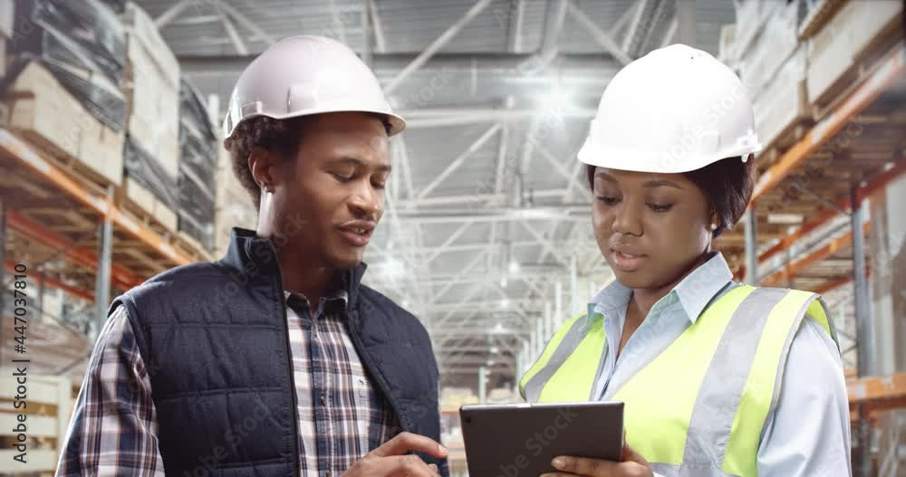 Two African-American workers, man and woman in protective helmets, recalculate cargo using tablet. Concept of teamwork in logistics company. Two African-American workers, man and woman in protective helmets, recalculate cargo using tablet. Concept of teamwork in logistics company.