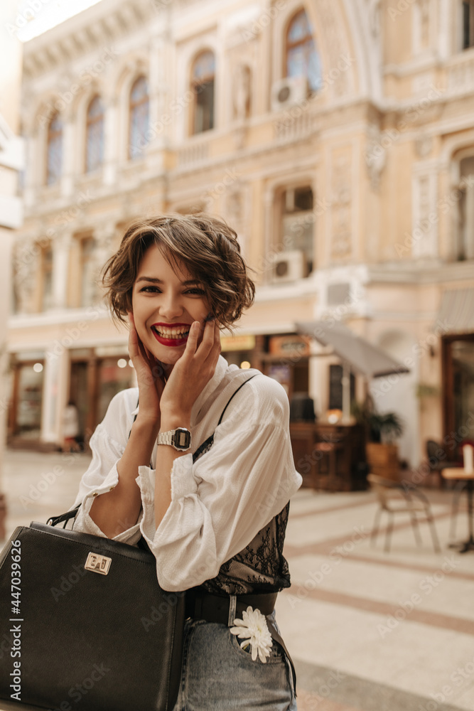Cheerful lady with wavy hair in blouse with black lace laughing at street. Trendy lady in jeans with handbag posing in city..