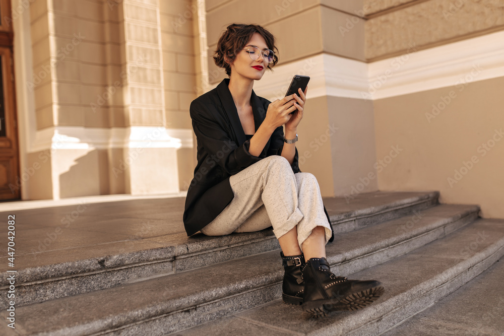 Wonderful lady in dark jacket and white pants holding phone outside. Short-haired woman in glasses sitting and smiling outdoors..