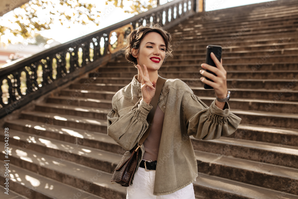 Fototapeta premium Cheerful girl in cool jacket and light jeans making selfie on backdrop of stairs. Young woman with wavy hair shows peace sign outside..