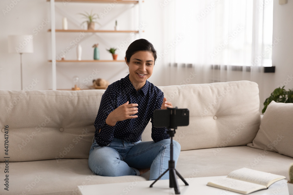 Smiling Indian millennial woman recording video on smartphone on tripod, sitting on couch at home, young female blogger influencer coach mentor shooting webinar or online training, talking on webcam