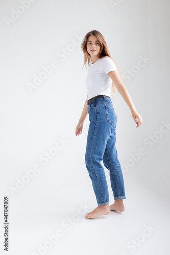 young attractive caucasian woman with long hair in t-shirt and blue jeans isolated on white studio background. skinny pretty female posing on cyclorama with bare feet. model tests of beautiful lady