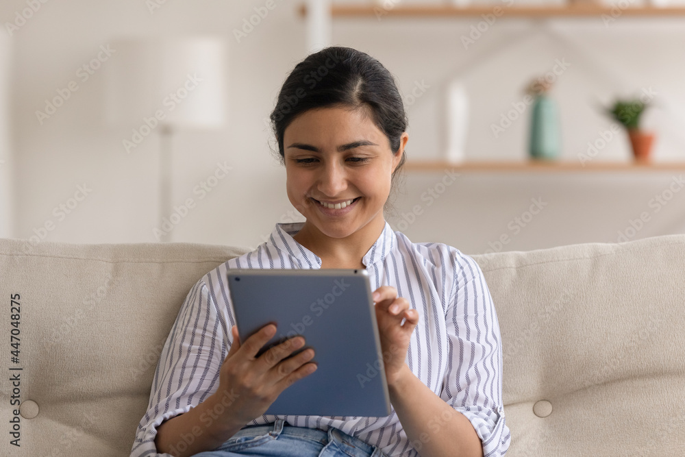 Close up smiling Indian woman using tablet, sitting on couch at home, happy positive young female looking at device screen, typing writing message, chatting in social network with friends, having fun Close up smiling Indian woman using tablet, sitting on couch at home, happy positive young female looking at device screen, typing writing message, chatting in social network with friends, having fun