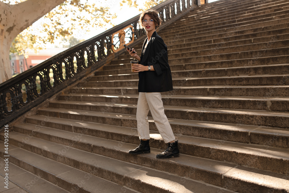 Full leght girl in boots, jacket and white trousers hold phone and cup of tes outside. Modern woman in eyeglasses posing on background of stairs..