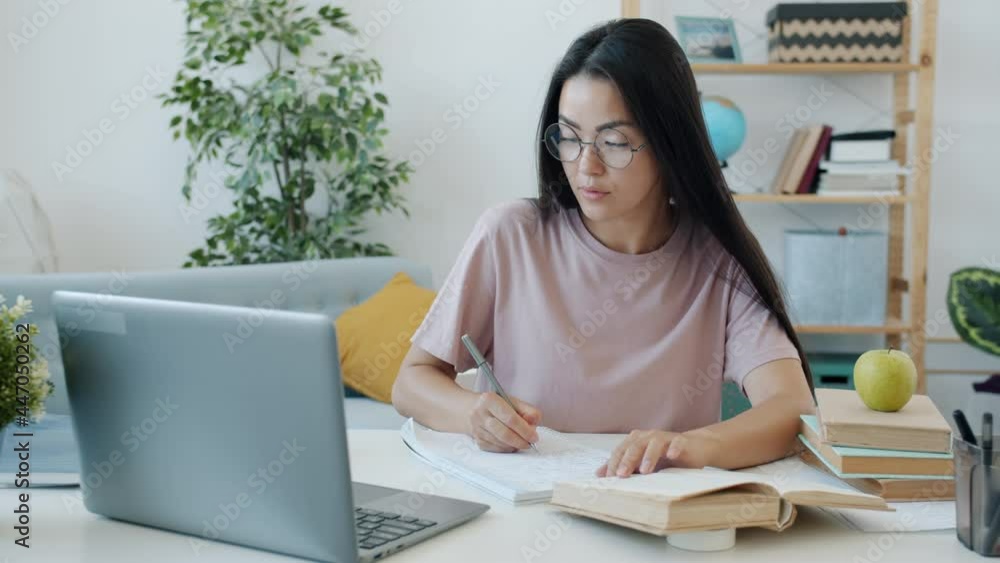 Slow motion of female Asian student doing homework using laptop and writing indoors at home. Distant education and modern technology concept. Slow motion of female Asian student doing homework using laptop and writing indoors at home. Distant education and modern technology concept.