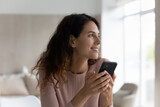 Dreamy millennial Latino woman use modern cellphone texting messaging online look in distance thinking planning. Smiling young Hispanic female client customer browse wireless internet on smartphone.