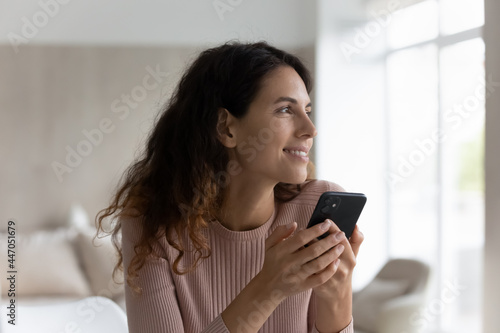 Dreamy millennial Latino woman use modern cellphone texting messaging online look in distance thinking planning. Smiling young Hispanic female client customer browse wireless internet on smartphone.