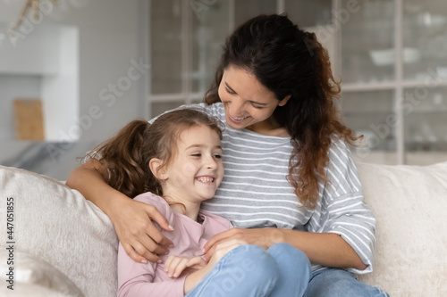Smiling young Latino mom and teen daughter relax on sofa at home have fun cuddling tickling. Happy Hispanic mother and teenage girl child rest on couch enjoy family lazy leisure weekend together.