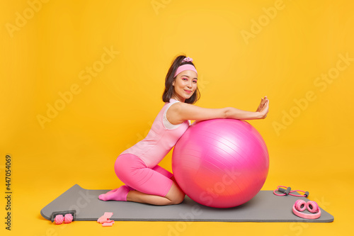 Sideways shot of pleased Asian woman lenas at fitness ball has satisfied expression dressed in activewear takes break after training at home likes gymnastics and aerobics poses on mat indoor