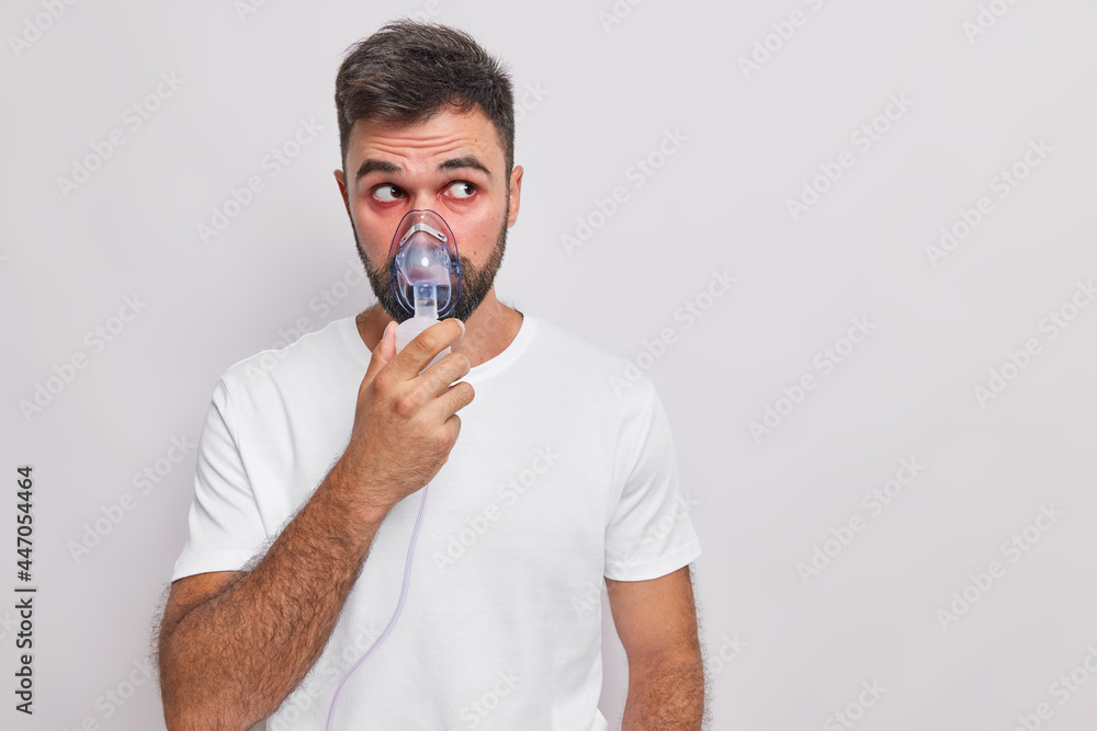 Health care and medical equipment concept. Unhealthy bearded man with red eyes wears oxygen mask has asthmatic attack does inhalation wears t shirt isolated over white background copy space. Health care and medical equipment concept. Unhealthy bearded man with red eyes wears oxygen mask has asthmatic attack does inhalation wears t shirt isolated over white background copy space.
