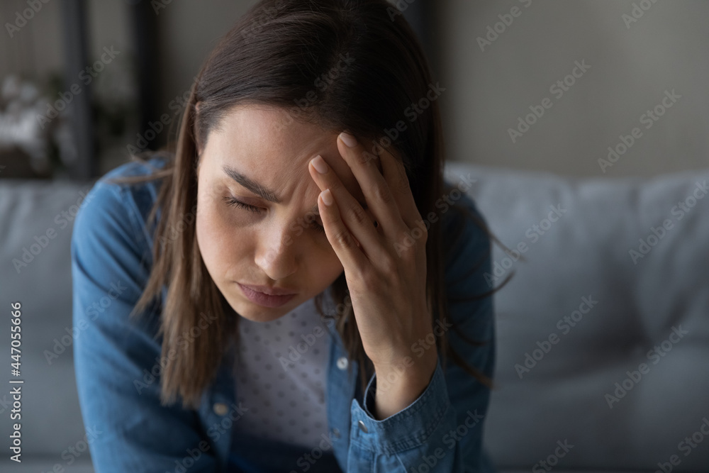 Close up of sad tired young woman sit on sofa, touch aching head having headache migraine looks upset troubled with life concerns feels stressed or sick suffers from personal problems, sorrow concept