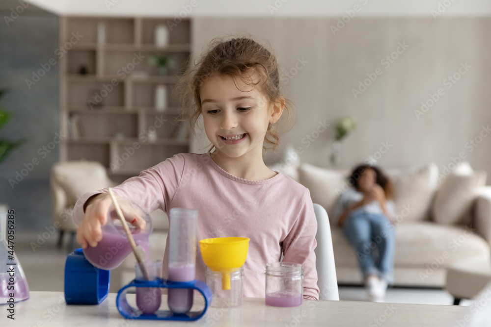Happy little teen ethnic girl child have fun engaged in chemistry lab activity at home alone. Smiling small biracial teenage kid play laboratory game on table, make experiments feel playful.
