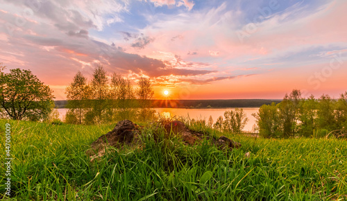 Scenic view at beautiful sunset on a shiny lake with old rough stone on the foreground  green grass  birch trees  golden sun rays  calm water  nice cloudy sky on a background  spring landscape