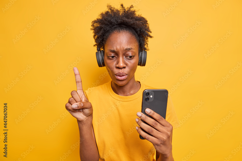 Serious attentive curly haired African American woman raises index finger looks at smartphone display listens music via headphones chooses song from playlist isolated over vivid yellow background Serious attentive curly haired African American woman raises index finger looks at smartphone display listens music via headphones chooses song from playlist isolated over vivid yellow background