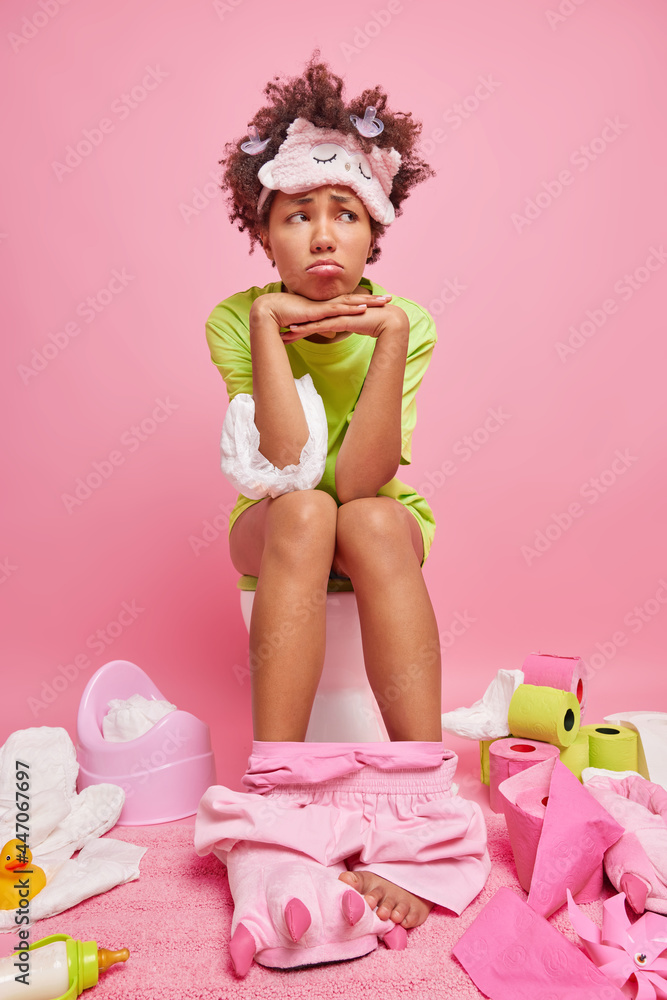 Fototapeta premium Vertical shot of displeased dark skinned ethnic woman with cury hair keeps hands under chin has unhappy expression while sitting on toilet bowl suffers from digestive disorder in lavatory room