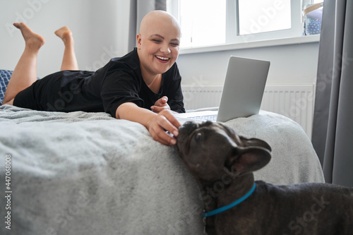 Bald woman feeding her dog while laying at the bed and working