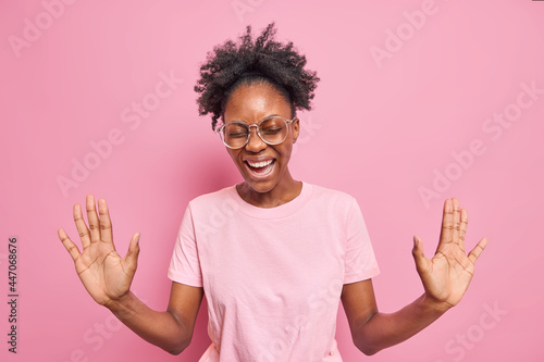 Authentic positive emotions and feelings concept. Cheerful dark skinned woman with natural curly hair keeps palms raised wears optical glasses casual t shirt isolated over pink studio background