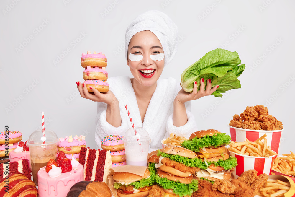 Happy lady holds donuts and vegetable faces difficult choice keeps to diet avoids eating harmful food applies patches under eyes after showering poses near table with various unhealthy products Happy lady holds donuts and vegetable faces difficult choice keeps to diet avoids eating harmful food applies patches under eyes after showering poses near table with various unhealthy products