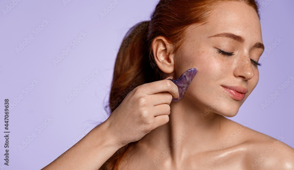 Fototapeta premium Beautiful redhead girl with clean healthy, glowing body, close eyes and smile pleased while using jade scraper on cheek, massaging facial, purple background