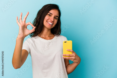 Young caucasian woman holding a mobile phone isolated on blue background cheerful and confident showing ok gesture.