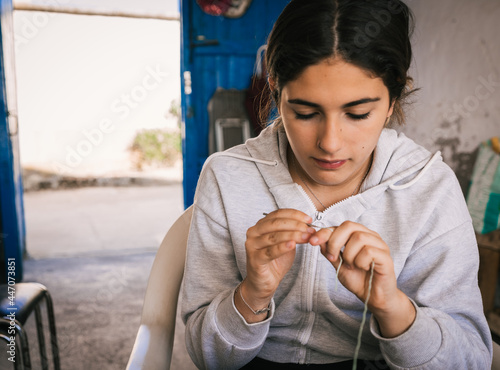 young brunette teenager in gray sweatshirt is making a bracelet with green yarn and crochet needle