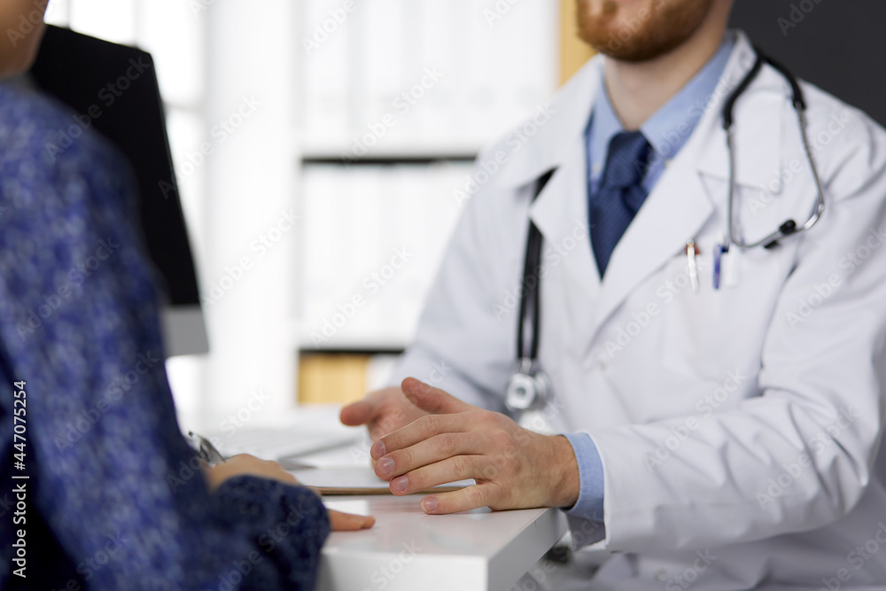 Unknown bearded doctor and patient woman discussing current health examination while sitting in sunny clinic, close-up. Medicine concept Unknown bearded doctor and patient woman discussing current health examination while sitting in sunny clinic, close-up. Medicine concept