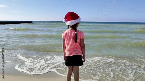 A 4-year-old girl in a Santa hat stands and looks at the sea