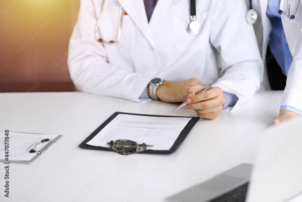 Fototapeta premium Group of unknown doctors are sitting at the desk and discussing problems or disease treatment in sunny hospital office. Covid 2019