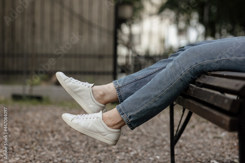 White shoes and jeans legs. Female legs in comfort casual urban sneakers. Close up photo of sexy women legs in white shoe sitting on the bench in the park. Sneakers mock-up. Walking.  photo