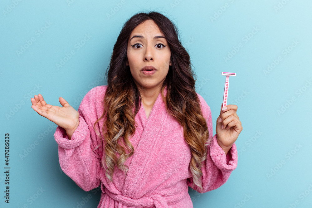 Young mexican woman wearing a bathrobe holding a knife isolated on blue background shrugs shoulders and open eyes confused. Young mexican woman wearing a bathrobe holding a knife isolated on blue background shrugs shoulders and open eyes confused.