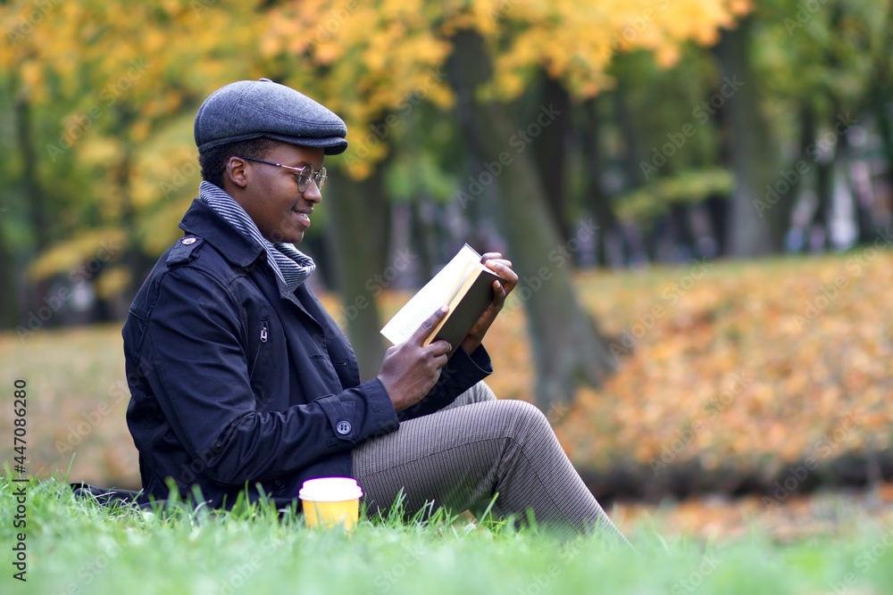 Happy black intelligent African Afro American guy, handsome stylish ethnic man reader is reading a book in glasses in a golden autumn park at fall, sitting on a grass with cup of coffee to go Happy black intelligent African Afro American guy, handsome stylish ethnic man reader is reading a book in glasses in a golden autumn park at fall, sitting on a grass with cup of coffee to go