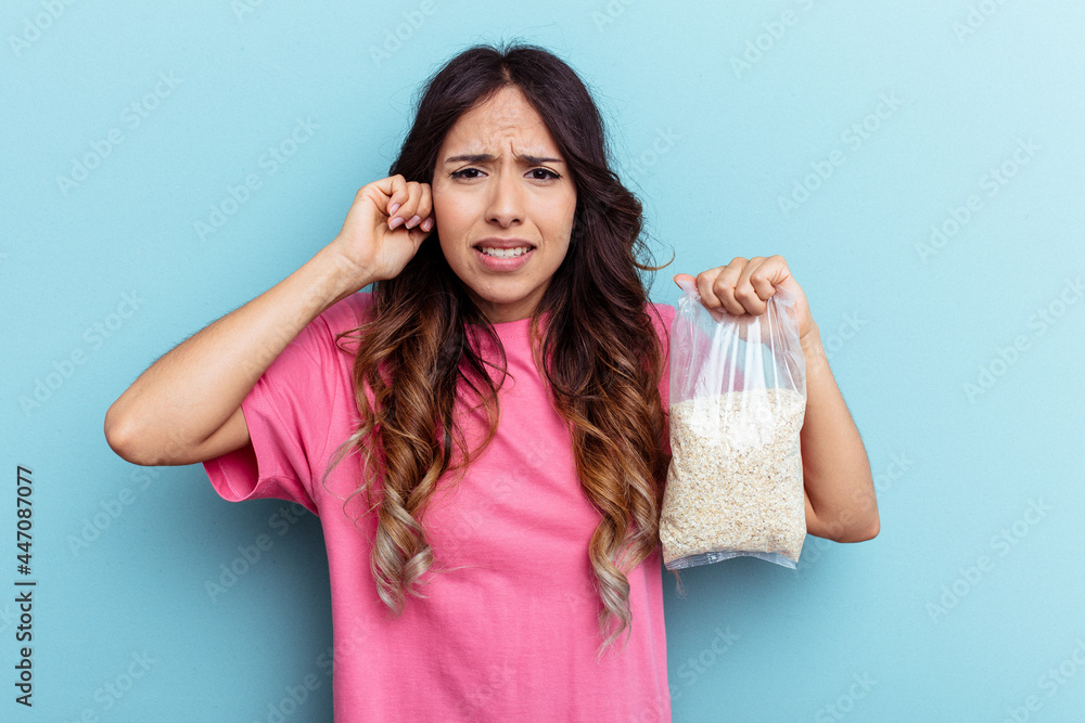 Young mixed race woman holding oatmeal isolated on blue background covering ears with hands. Young mixed race woman holding oatmeal isolated on blue background covering ears with hands.