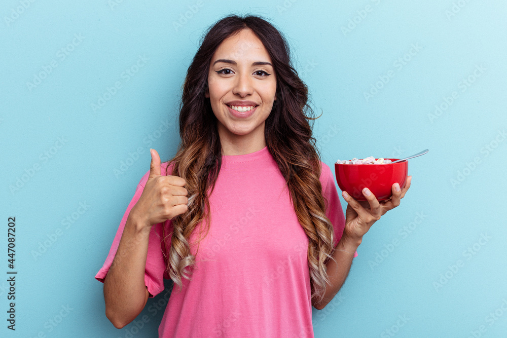 Young mixed race woman holding cereals bowl isolated on blue background smiling and raising thumb up Young mixed race woman holding cereals bowl isolated on blue background smiling and raising thumb up
