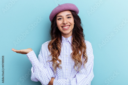 Young mexican woman isolated on blue background showing a copy space on a palm and holding another hand on waist.