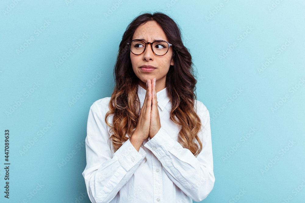 Young mexican woman isolated on blue background praying, showing devotion, religious person looking for divine inspiration. Young mexican woman isolated on blue background praying, showing devotion, religious person looking for divine inspiration.