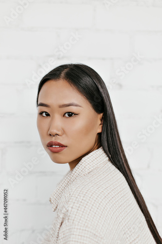 Portrait of brunette brown-eyed woman poses on white brick wall background. Attractive Asian lady looks into camera.