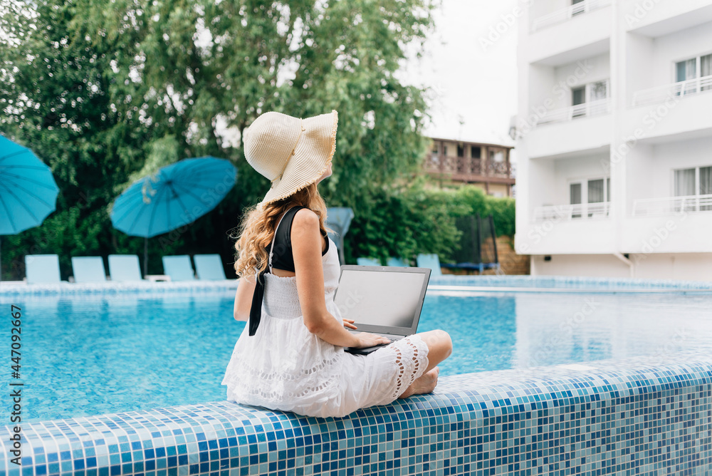 Fototapeta premium At a resort hotel near the pool sits a young happy woman in a straw hat with a laptop. technology concept and remote work.