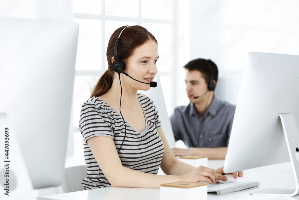 Casual dressed young woman using headset and computer while talking with customers online. Group of operators at work. Call center, business concept Casual dressed young woman using headset and computer while talking with customers online. Group of operators at work. Call center, business concept