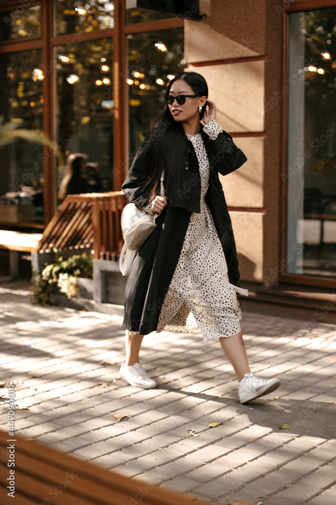 Fototapeta premium Cheerful brunette woman in midi polka dot dress and black stylish trench moves outside. Attractive lady in sunglasses walks on street.