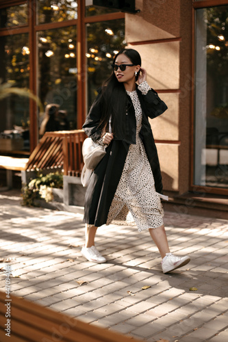 Cheerful brunette woman in midi polka dot dress and black stylish trench moves outside. Attractive lady in sunglasses walks on street.