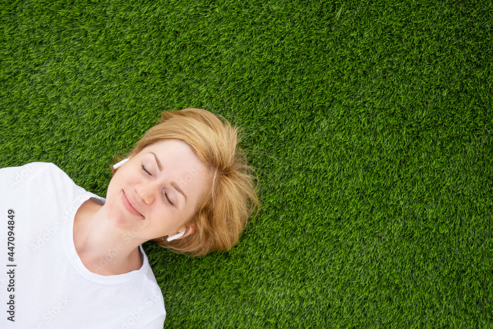 A young Caucasian girl in a white T-shirt and headphones lies on an artificial lawn with closed eyes. The concept of listening to music, podcasts. Minimalism, copy space, top view. A young Caucasian girl in a white T-shirt and headphones lies on an artificial lawn with closed eyes. The concept of listening to music, podcasts. Minimalism, copy space, top view.